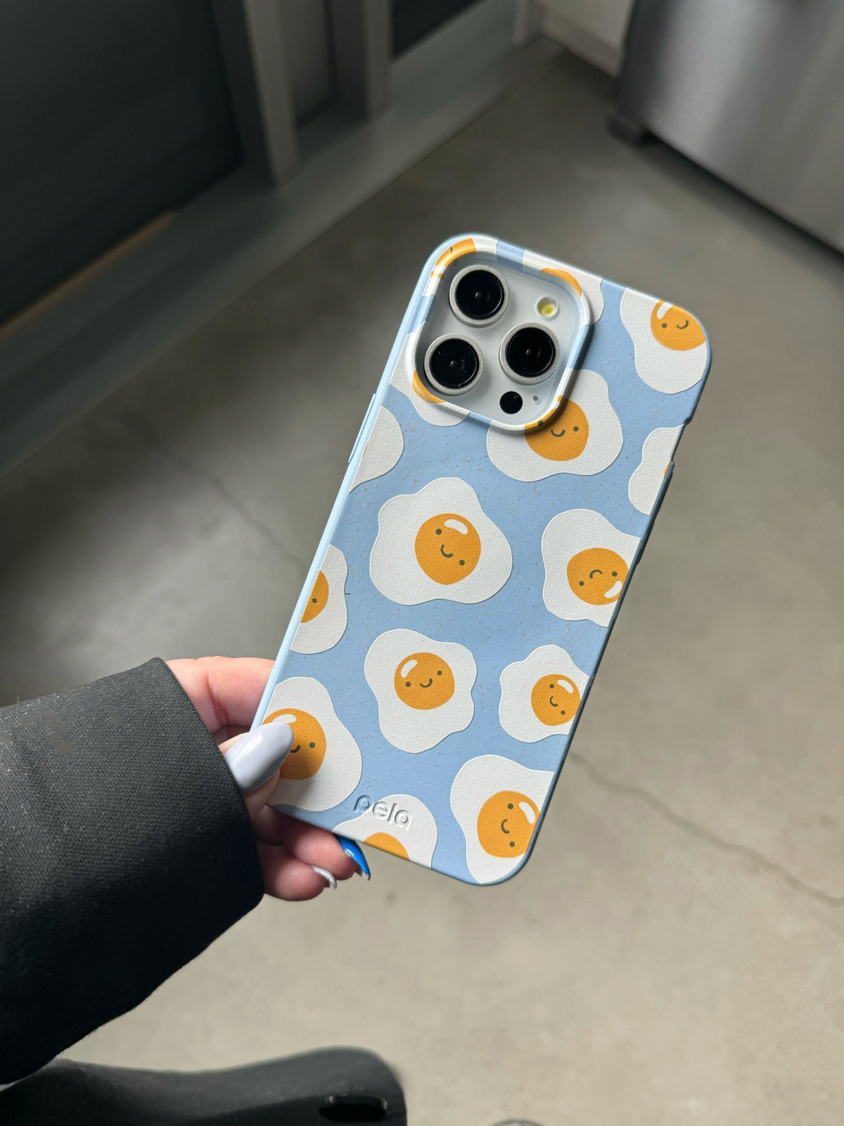 A hand holds Pela's Powder Blue Sunny Side Up Phone Case from their Breakfast Foods Collection. The case features a playful design with smiling fried eggs on a light blue background. The person holding the case has light-colored nails and is wearing a black sleeve.