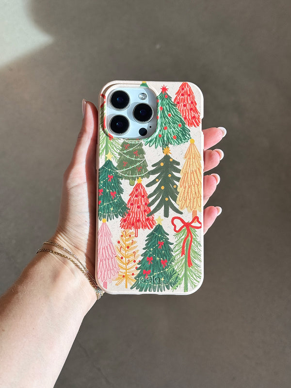 A hand holds the Pela Festive Trees Phone Case, featuring a colorful pattern of illustrated Christmas trees in shades of green, red, pink, and gold on a light beige background.