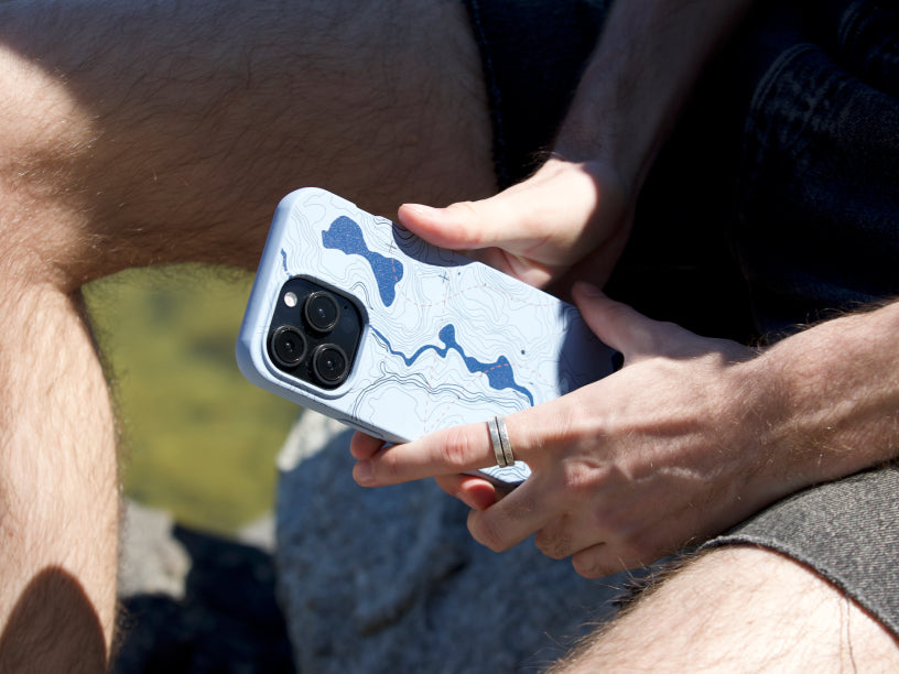 A person holding the Pela Contour Lines Phone Case, which features a topographic map design in shades of blue. The phone case is being held outdoors, with the person seated on rocks, highlighting the case’s adventure-ready aesthetic.