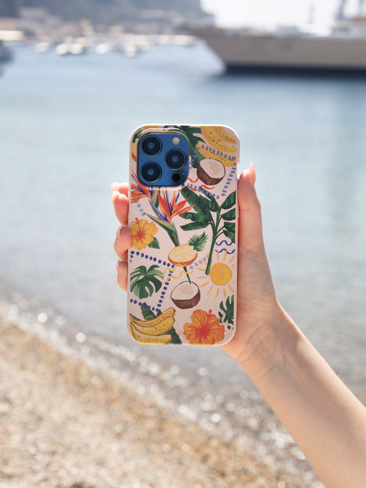 The phone case featured in the image is the Pela Tropics Phone Case from the Summer Travel Collection, held in a hand against a scenic seaside background with boats and a rocky shoreline in the distance. The case design includes tropical elements such as bananas, coconuts, and vibrant flowers.