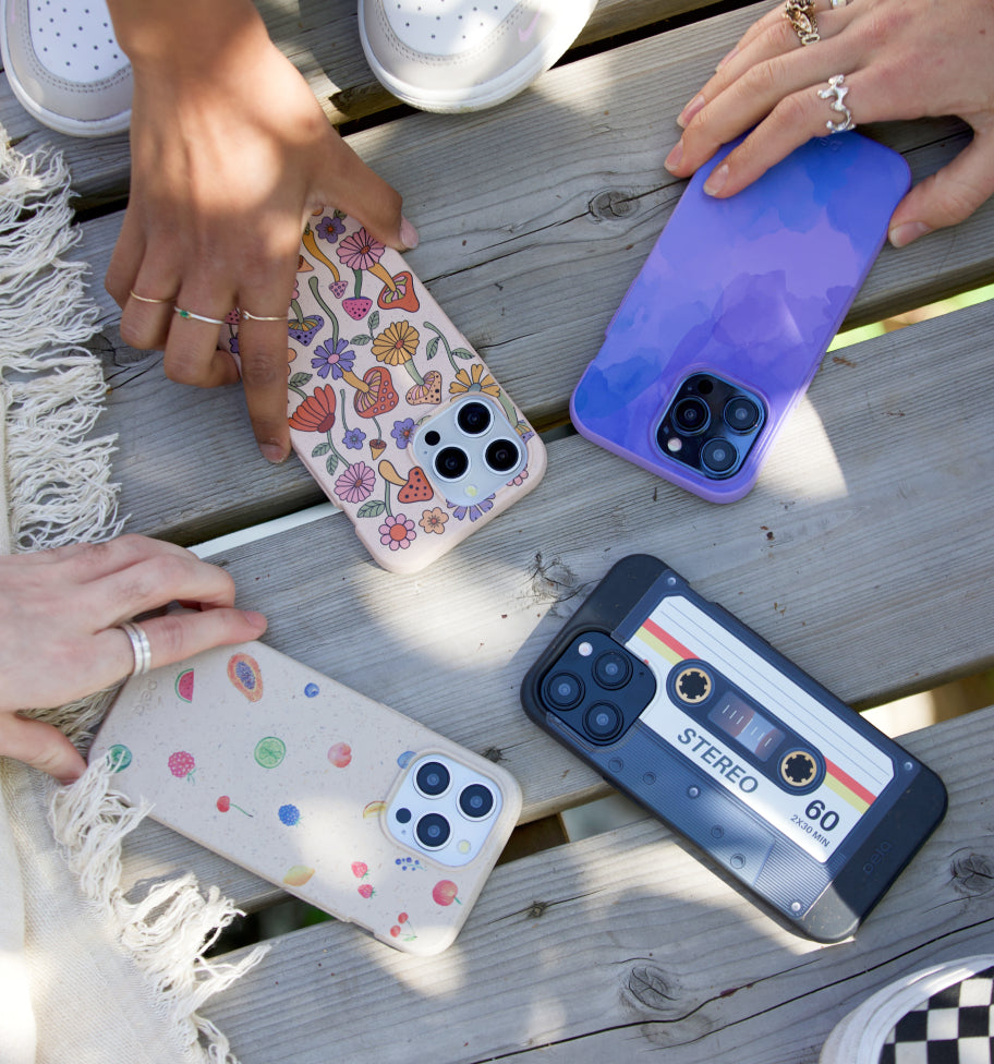 A group of Pela phone cases placed on a wooden surface, with hands reaching towards them. The designs include a colorful mushroom and floral pattern, a solid purple gradient, a retro cassette tape design, and a playful fruit motif. The scene captures the cases’ eco-friendly charm while showing off their unique, vibrant artwork.